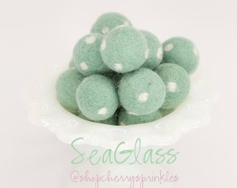 May include: A white bowl filled with light green felt balls with white polka dots. The bowl has a scalloped edge. The text "SeaGlass" and "@shopcherryssprinkles" is written below the bowl.