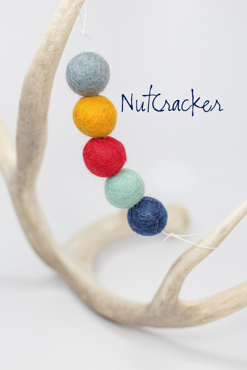 May include: A garland of felt balls in shades of blue, yellow, red, and green, hanging from a white antler. The garland is labeled "Nutcracker".
