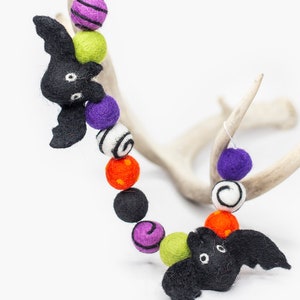 May include: A Halloween garland with black felt bats, green, purple, orange, and white felt balls with various patterns. The garland is draped over a white antler.