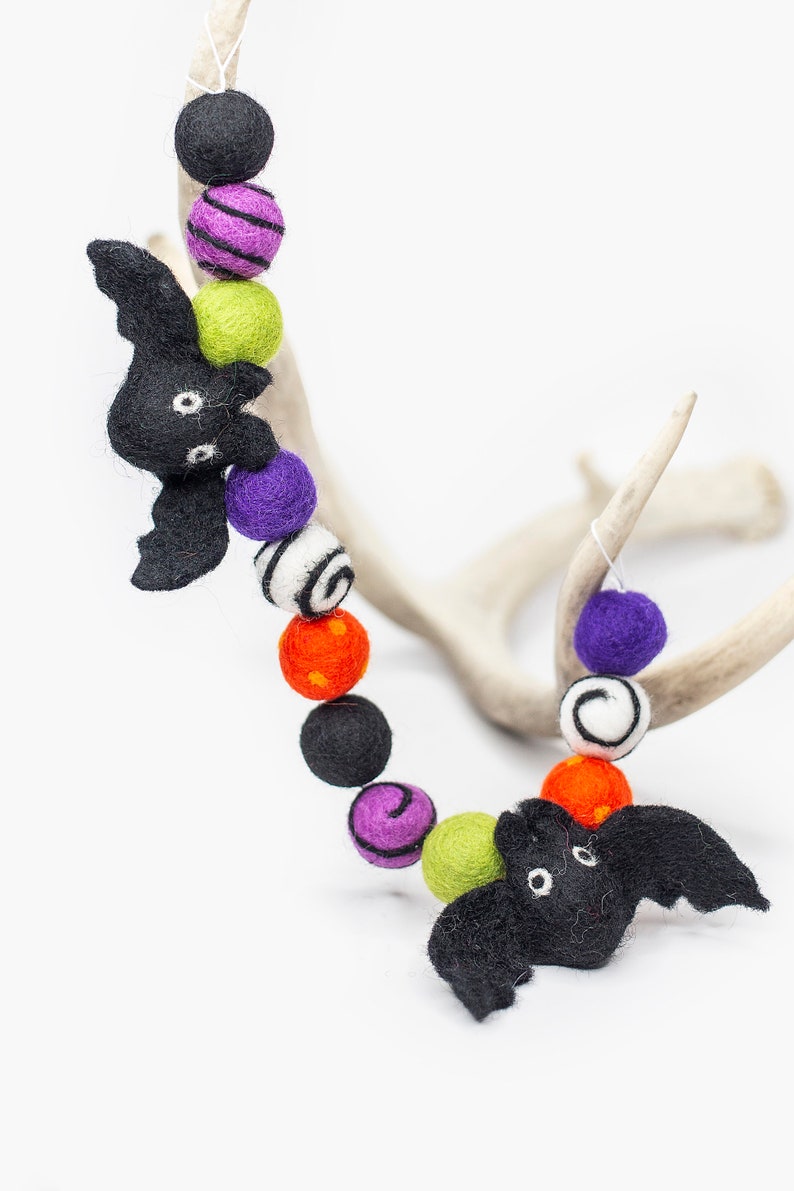 May include: A Halloween garland with black felt bats, green, purple, orange, and white felt balls with various patterns. The garland is draped over a white antler.