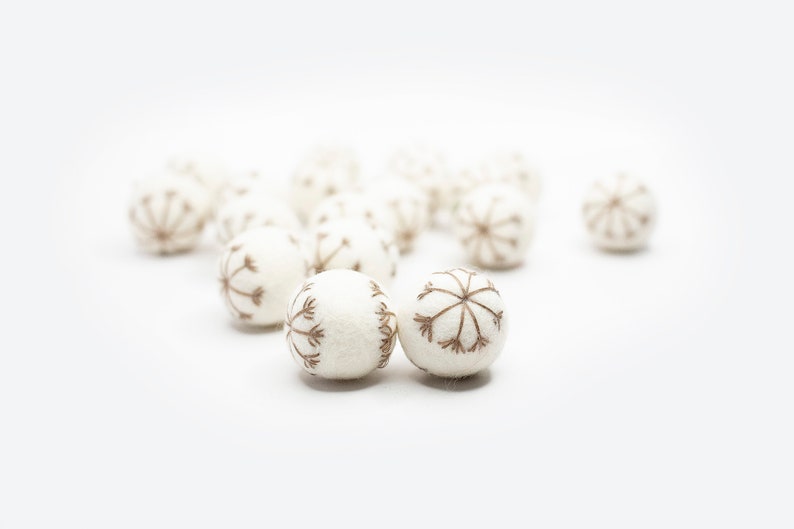 Puede incluir: Un grupo de bolas de fieltro blanco con dise&ntilde;os de copo de nieve bordados en marr&oacute;n. Las bolas est&aacute;n esparcidas sobre una superficie blanca.