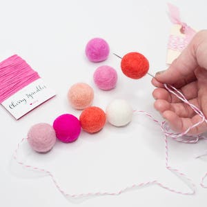 May include: A collection of colorful felt balls in shades of pink, red, and white, strung on a white and pink twine. A needle is used to thread a red felt ball. The words "Cherry Sprinkles" are printed on a small card.