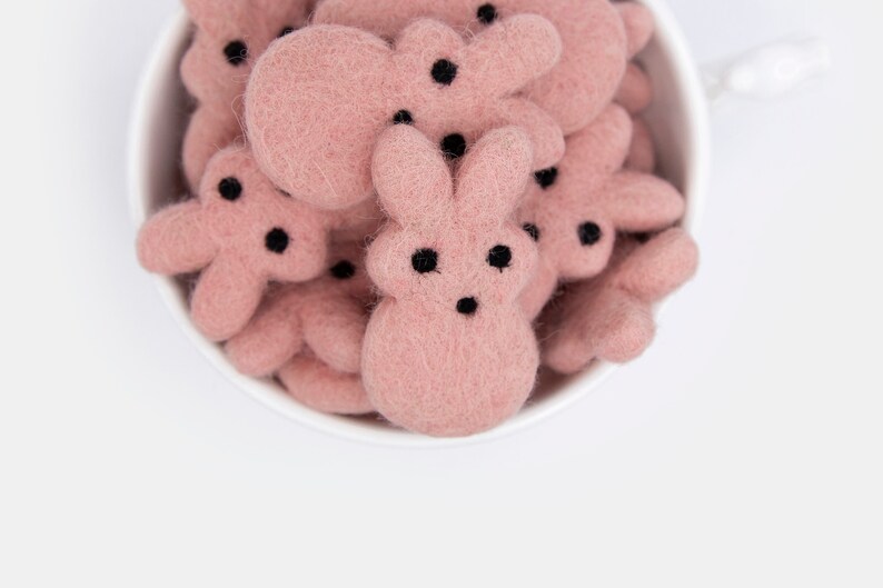 May include: A white ceramic bowl filled with pink felt bunny shapes. The bunnies have black eyes and noses.