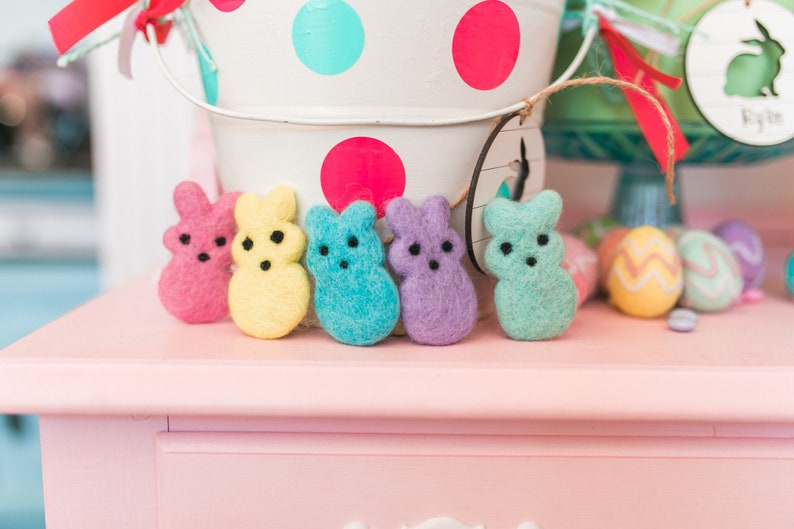 May include: Six felt bunny shaped candies in various pastel colors, including pink, yellow, blue, turquoise, purple, and green. The candies are arranged in a row on a pink surface.