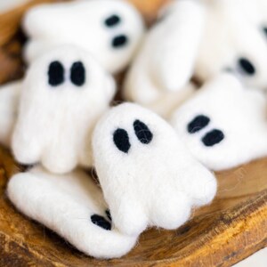 May include: A bowl filled with white felt ghost ornaments with black felt eyes. The ghosts are arranged in a cluster, with some overlapping.