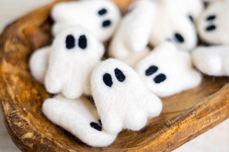 May include: A bowl filled with white felt ghost ornaments with black felt eyes. The ghosts are arranged in a cluster, with some overlapping.