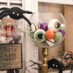 May include: A glass jar filled with colorful felt eyeballs and a black spider on top. The jar has a label that reads "Original Wartcap Powder Slug & Jiggers Apothecary For external use only".