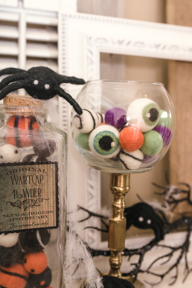 May include: A glass jar filled with colorful felt eyeballs and a black spider on top. The jar has a label that reads "Original Wartcap Powder Slug & Jiggers Apothecary For external use only".