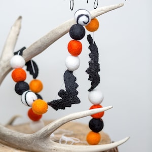 May include: A Halloween garland featuring black felt bats and colorful felt balls in orange, black, and white. The garland is displayed on a deer antler and a wooden base. The text "Spooky Bats" is written in a script font.