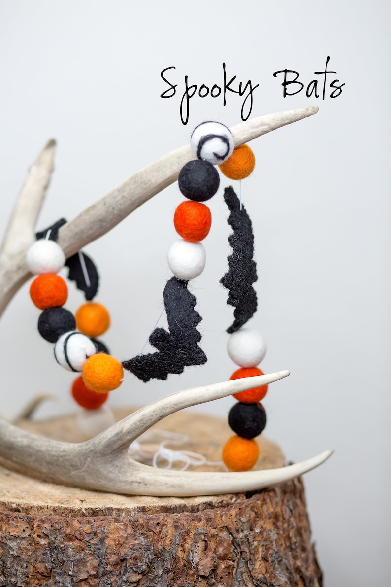 May include: A Halloween garland featuring black felt bats and colorful felt balls in orange, black, and white. The garland is displayed on a deer antler and a wooden base. The text "Spooky Bats" is written in a script font.