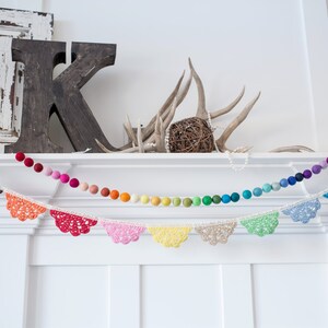 May include: A decorative garland featuring a string of colorful felt balls and a separate string of crochet lace pennants. The felt balls transition through the colors of the rainbow. The pennants are in various colors.