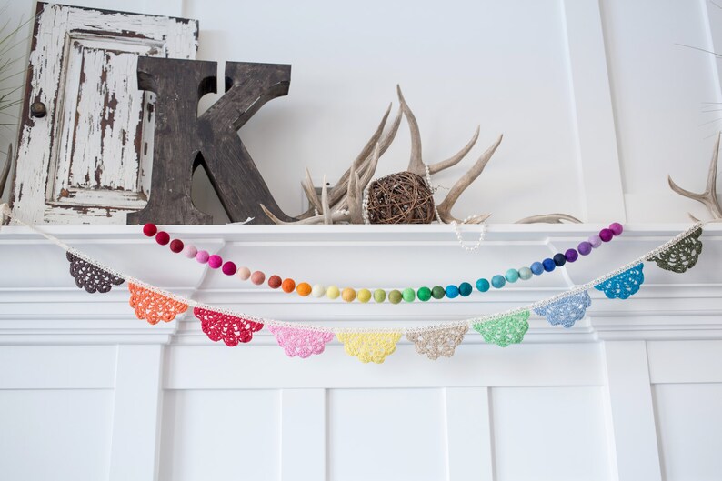May include: A decorative garland featuring a string of colorful felt balls and a separate string of crochet lace pennants. The felt balls transition through the colors of the rainbow. The pennants are in various colors.