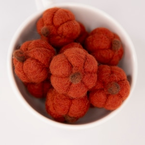 May include: A white ceramic cup filled with six small, orange felt pumpkins. The pumpkins are arranged in a cluster, with some overlapping.