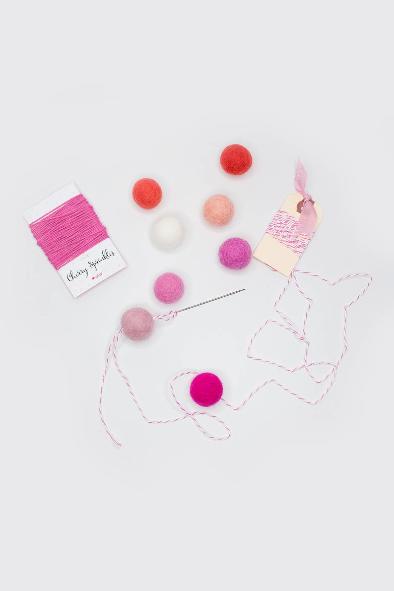 May include: A pink and white string with a small tag that says "Cherry Sprinkles" and a pink heart. There are also several pink, white, and orange felt balls and a needle.
