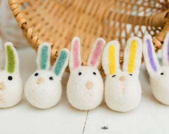 May include: Five white felt bunny ornaments with colorful ears. The bunnies are lined up in a row on a white surface. The bunnies have black eyes and a brown nose.