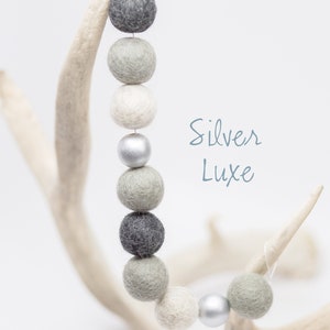 Pu&ograve; includere: Una ghirlanda di palline di feltro in tonalit&agrave; di grigio e bianco con un perlino argentato, appesa a un palco di cervo bianco. Il testo "Silver Luxe" &egrave; scritto in corsivo blu.