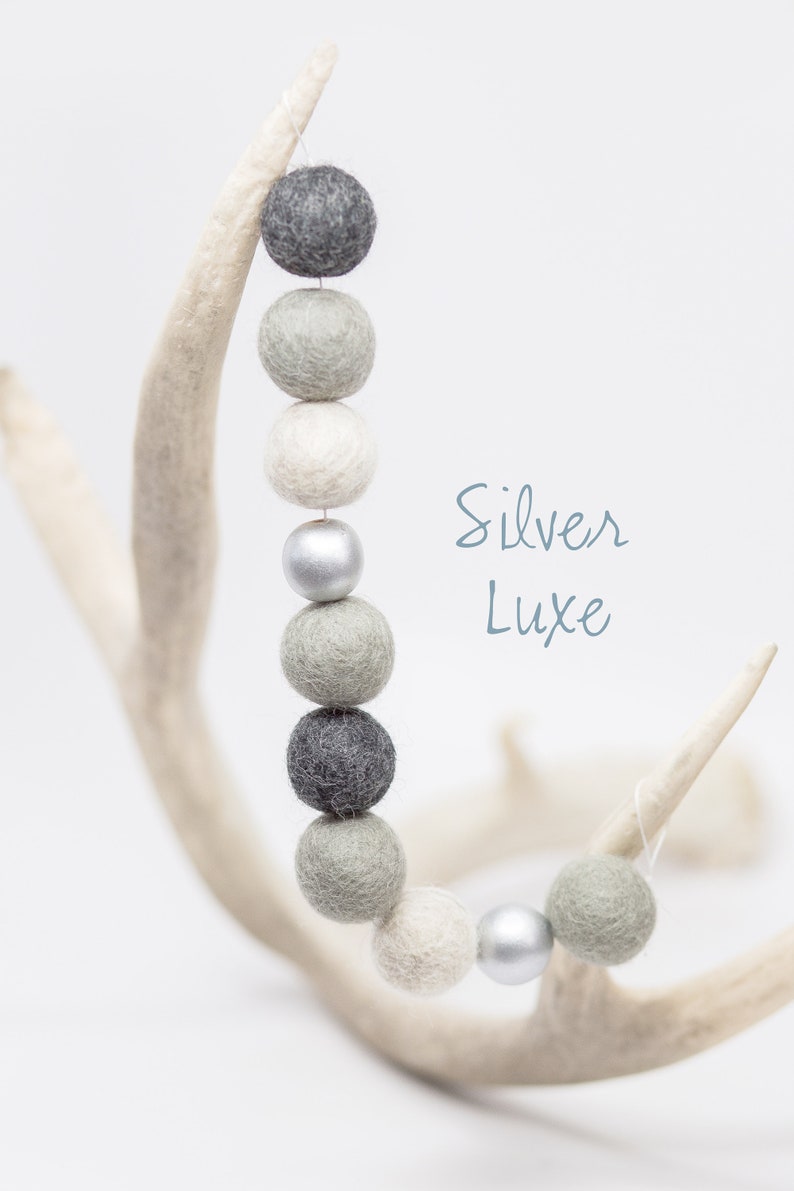 Pu&ograve; includere: Una ghirlanda di palline di feltro in tonalit&agrave; di grigio e bianco con un perlino argentato, appesa a un palco di cervo bianco. Il testo "Silver Luxe" &egrave; scritto in corsivo blu.