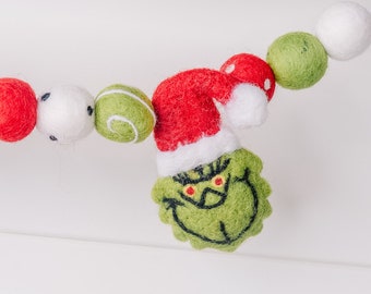 May include: A festive Christmas garland featuring red, white, and green felt balls. The garland includes a green Grinch character wearing a red Santa hat. The Grinch has yellow eyes and a stitched black mouth. The garland is strung on a white string.