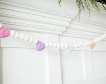 May include: A garland of white felt balls with pink, purple, and light green felt hearts strung between them. The garland is hanging on a white wall.
