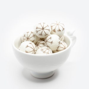 Puede incluir: Un taz&oacute;n de cer&aacute;mica blanco lleno de bolas de fieltro blancas decoradas con dise&ntilde;os de copos de nieve marrones.