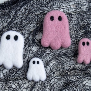 May include: Four felt ghost ornaments in pink and white. The ghosts are arranged on a black mesh background.