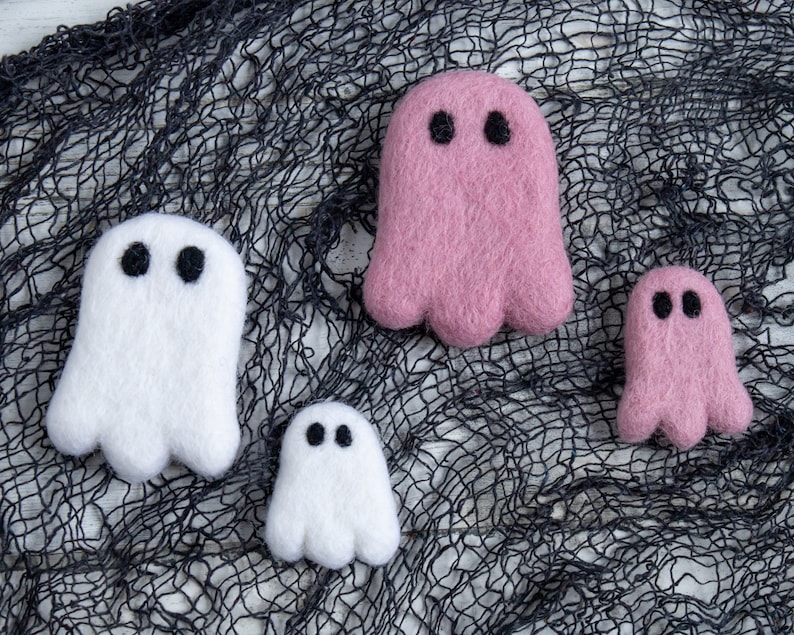 May include: Four felt ghost ornaments in pink and white. The ghosts are arranged on a black mesh background.