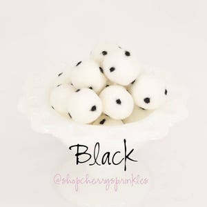 May include: A white bowl filled with white pom poms with black polka dots. The text "Black" is written below the bowl. The text "@shopcherryssprinkles" is written below the word "Black".