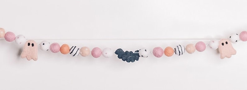 May include: A pink and white felt garland with ghosts and bats. The garland is strung on a white string and has pink, white, and orange felt balls.