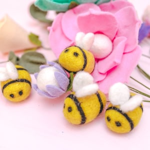 May include: Five felt bumblebees with black stripes and white wings are arranged on a pink surface with a pink felt flower and a purple felt flower with a white center.