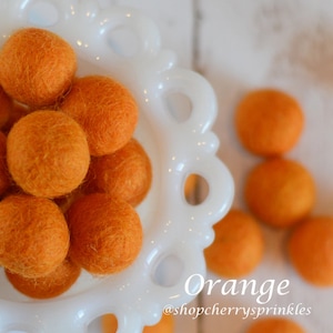 May include: A white ceramic bowl filled with orange felt balls. The bowl has a decorative edge and is sitting on a white wooden surface. The text "Orange" and "@shopcherry sprinkles" is visible below the bowl.