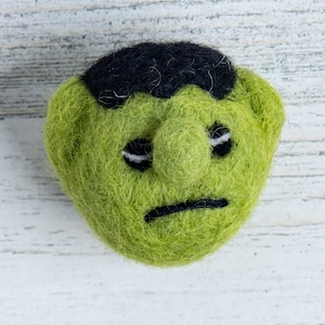 Pu&ograve; includere: Mostro di feltro verde con capelli di feltro nero e un'espressione triste.