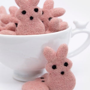 May include: A white teacup filled with pink felt bunny shapes. Two more pink felt bunnies are sitting on a white surface in front of the teacup.