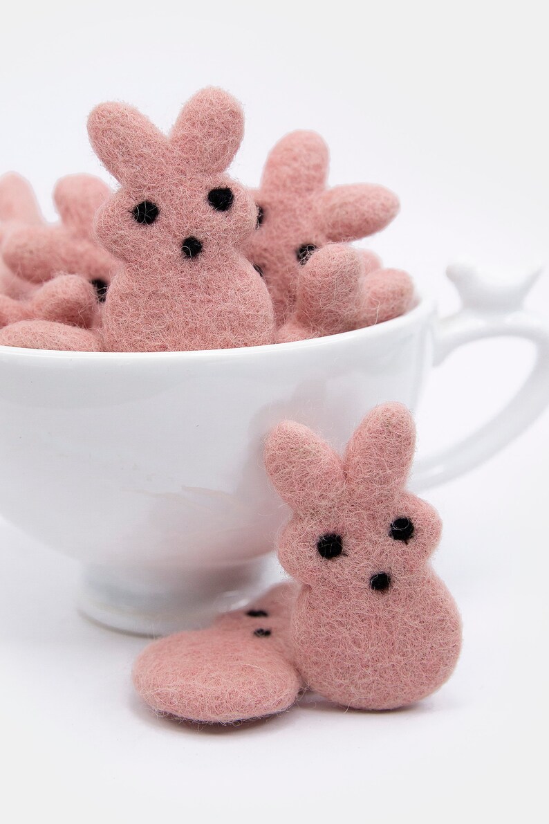 May include: A white teacup filled with pink felt bunny shapes. Two more pink felt bunnies are sitting on a white surface in front of the teacup.