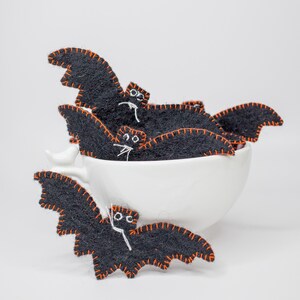 May include: A collection of black felt bat decorations with orange stitching and white facial details. Some bats are inside a white teacup, while others are displayed in front. These Halloween-themed decorations are handmade.