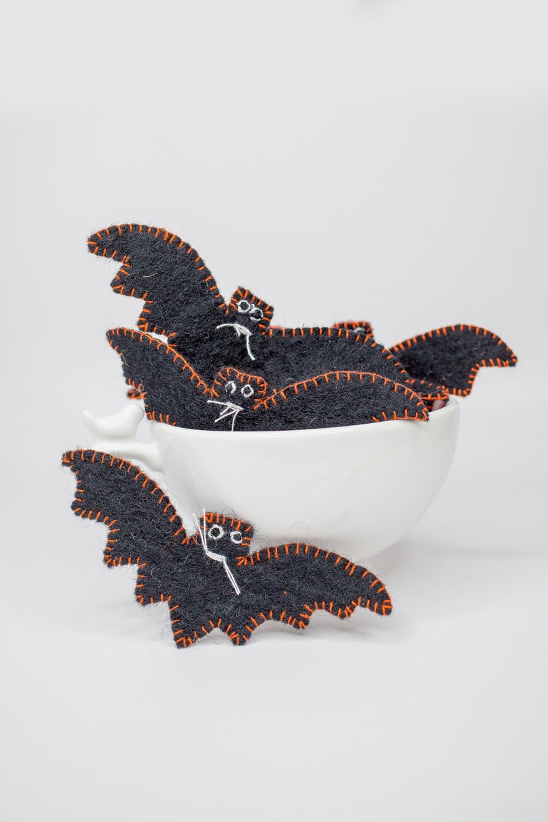 May include: A collection of black felt bat decorations with orange stitching and white facial details. Some bats are inside a white teacup, while others are displayed in front. These Halloween-themed decorations are handmade.
