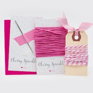 May include: A pink and white twine with a pink ribbon and a needle and thread on a white background. The twine is labeled "Cherry Sprinkle" and "shop etsy".
