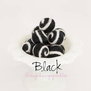 May include: A white bowl filled with black and white felt balls. The balls have a swirl pattern. The text "Black" and "@shopcherryssprinkles" is below the bowl.