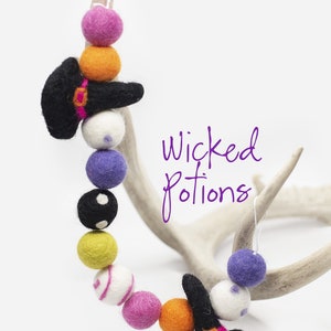 May include: A felt garland with black witch hats, orange, pink, white, purple, yellow, and black and white polka dot felt balls. The garland is hanging on a white antler. The text "Wicked Potions" is written in purple on a white background.