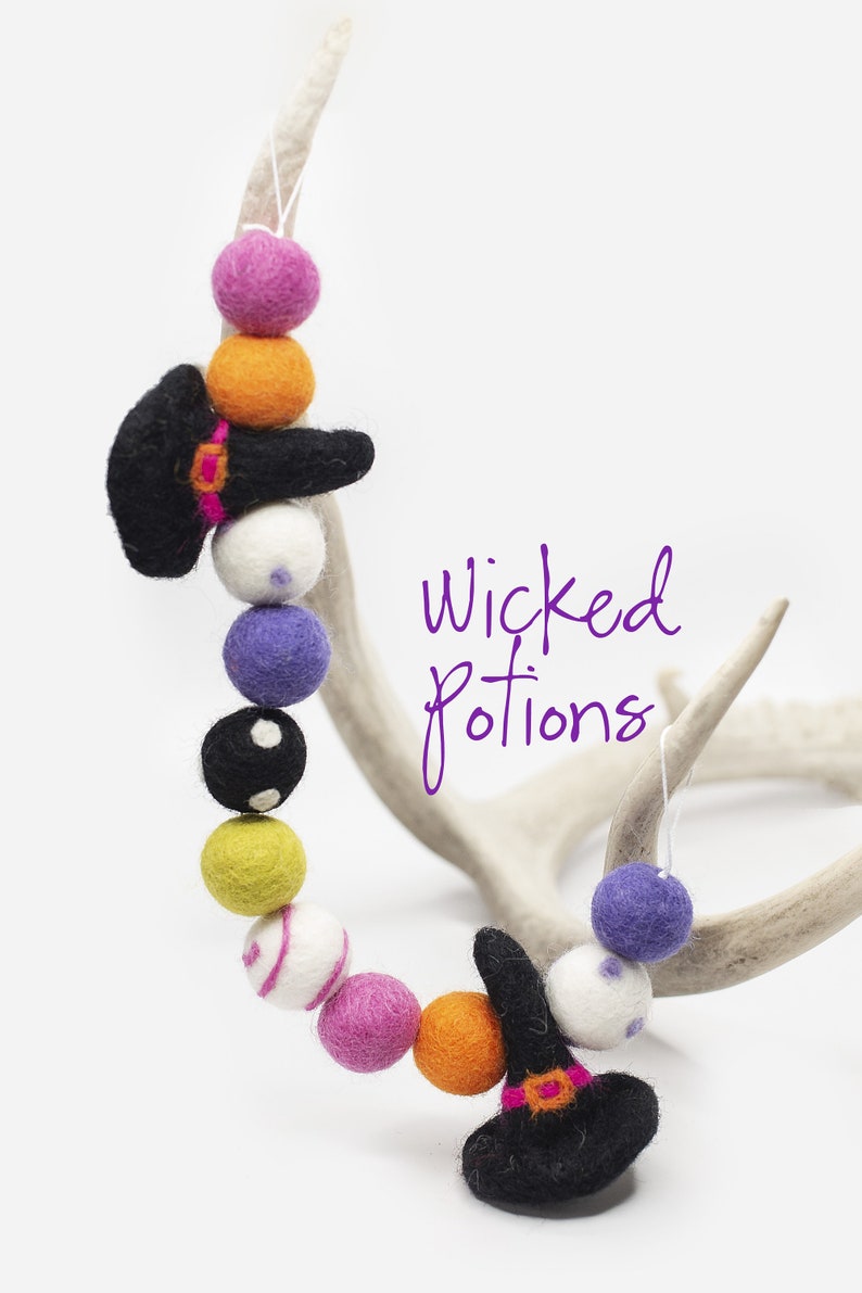 May include: A felt garland with black witch hats, orange, pink, white, purple, yellow, and black and white polka dot felt balls. The garland is hanging on a white antler. The text "Wicked Potions" is written in purple on a white background.