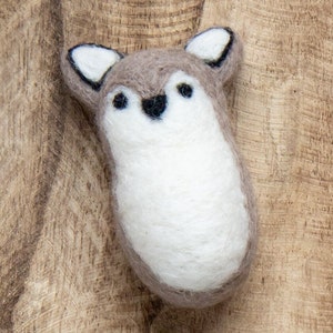 May include: A small, brown and white felt animal with black eyes and a black nose. The animal is shaped like a fox or a wolf and is sitting on a wooden surface.