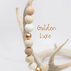 Pu&ograve; includere: Una ghirlanda di palline di feltro beige, crema e bianco con dettagli dorati. La ghirlanda &egrave; appesa a un paio di corna. Il testo "Golden Luxe" &egrave; scritto in lettere dorate.