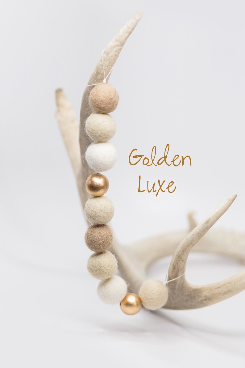 Pu&ograve; includere: Una ghirlanda di palline di feltro beige, crema e bianco con dettagli dorati. La ghirlanda &egrave; appesa a un paio di corna. Il testo "Golden Luxe" &egrave; scritto in lettere dorate.
