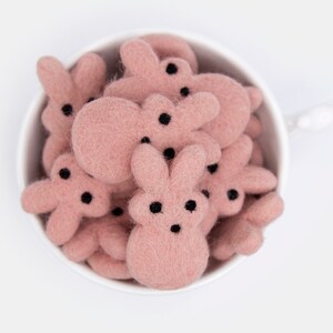 May include: A white ceramic cup filled with pink felt bunny shapes. The bunnies have black eyes and noses.