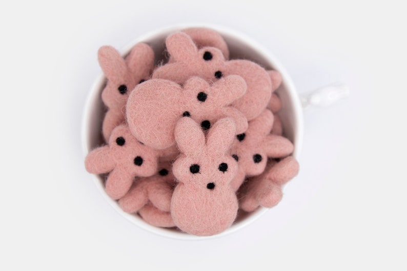 May include: A white ceramic cup filled with pink felt bunny shapes. The bunnies have black eyes and noses.