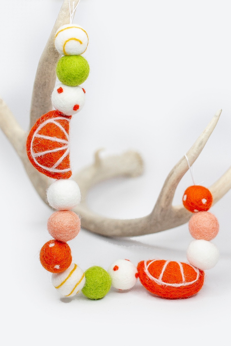 May include: A garland of felt balls in orange, white, green, and peach colors. The garland features orange felt half-circles with white stitching, resembling citrus fruit slices.