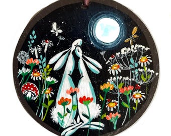 Könnte beinhalten: Runder, handbemalter Holzschmuck mit einer skurrilen Szene von zwei weißen Hasen, die sich unter einem Vollmond umarmen. Der dunkle Hintergrund ist mit Sternen übersät und von bunten Blumen, Schmetterlingen und einem Marienkäfer umgeben. Ein dekoratives Stück mit rustikalem Charme.