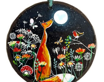 Könnte beinhalten: Runder, handbemalter Holzschmuck mit einem Fuchs, der den Mond in einem sternenklaren Nachthimmel betrachtet. Bunte Blumen, ein Vogel und Insekten umgeben den Fuchs. Der Schmuck hat eine türkisfarbene Schnur zum Aufhängen.
