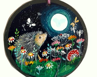 Könnte beinhalten: Rundes, handgemaltes Kunstwerk mit einem Igel, der einen Vollmond in einem sternenklaren Nachthimmel betrachtet. Bunte Wildblumen und ein roter Pilz mit weißen Punkten sind im Vordergrund. Eine rosa Schnur ist zum Aufhängen befestigt.