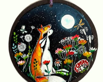 Könnte beinhalten: Ein handbemalter, runder Holzschmuck mit einem Fuchs und einem Hasen, die sich unter einem sternenklaren Nachthimmel mit Vollmond umarmen. Die Szene umfasst bunte Wildblumen, Löwenzahnpflanzen und eine Libelle. Eine lila Schnur ist zum Aufhängen befestigt.