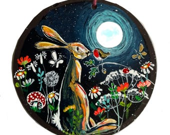 Könnte beinhalten: Runder Holzschmuck mit einer skurrilen Illustration eines Hasen und eines Rotkehlchens unter einem Vollmond. Das Kunstwerk umfasst bunte Blumen, Schmetterlinge und einen Pilz vor einem dunkelblauen Nachthimmel. Eine bordeauxfarbene Schnur ist zum Aufhängen befestigt.